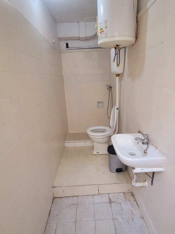 Baño alargado con azulejo blanco, termotanque eléctrico colgado con sus conexiones a la vista, inodoro, lavamanos mural y zona de ducha al fondo.